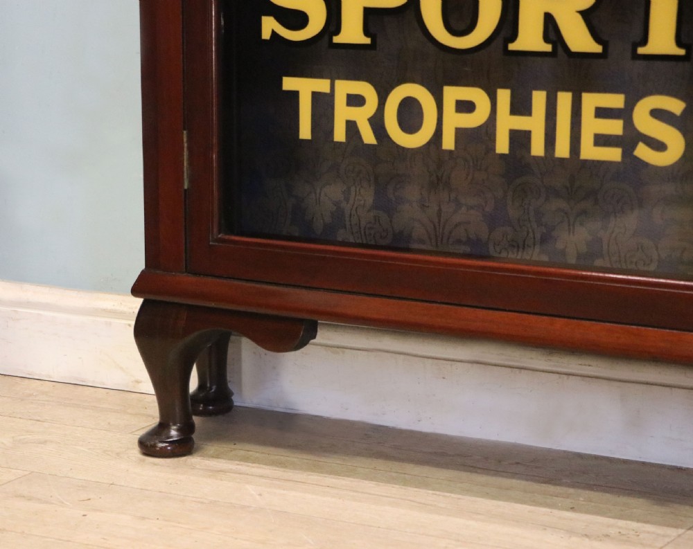 A late nineteenth-century dark mahogany display cabinet featuring two glazed doors adorned with hand-painted gold advertising typography, resting on short cabriole legs. View XXIV