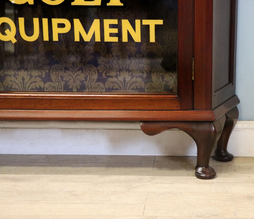 A late nineteenth-century dark mahogany display cabinet featuring two glazed doors adorned with hand-painted gold advertising typography, resting on short cabriole legs. View XXVI
