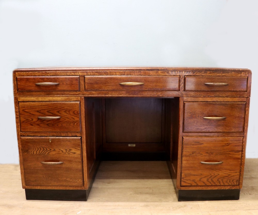 A twin-pedestal mid-century oak office desk featuring seven drawers with brass pull handles and a dark plinth base.