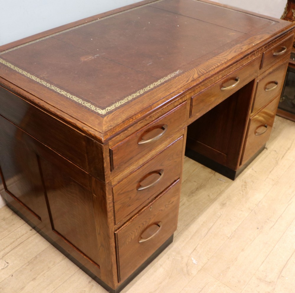 A twin-pedestal mid-century oak office desk featuring seven drawers with brass pull handles and a dark plinth base. View V