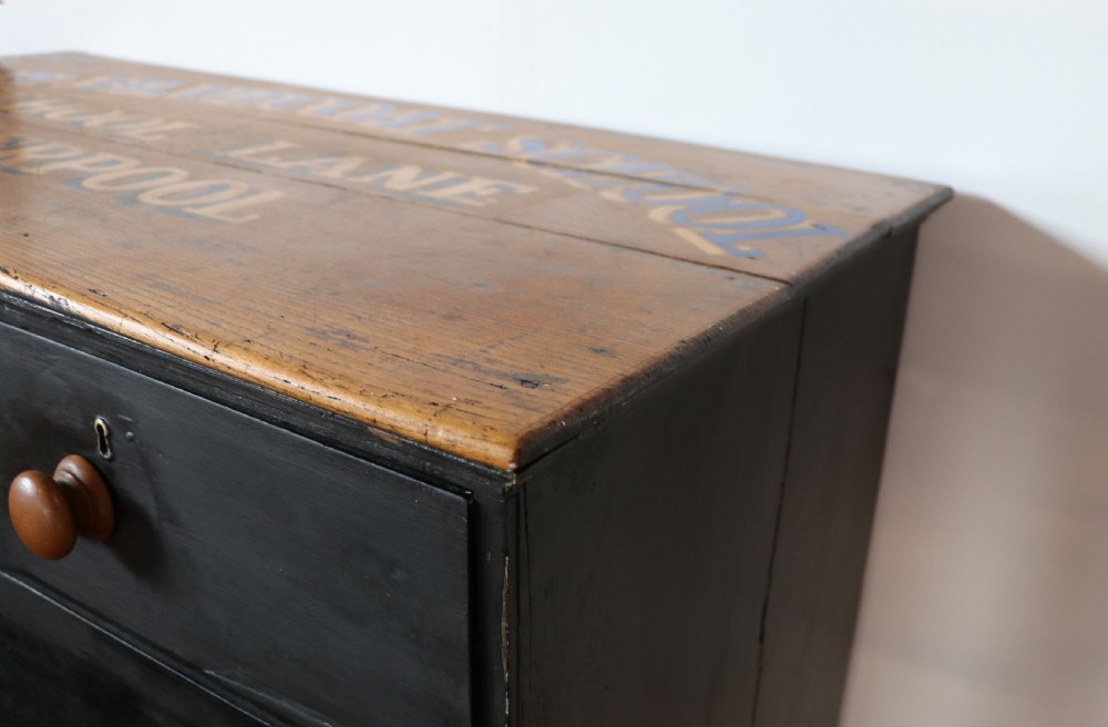 A Victorian black painted pine chest of drawers featuring a natural timber top with hand-painted lettering and turned wooden knobs. View XV