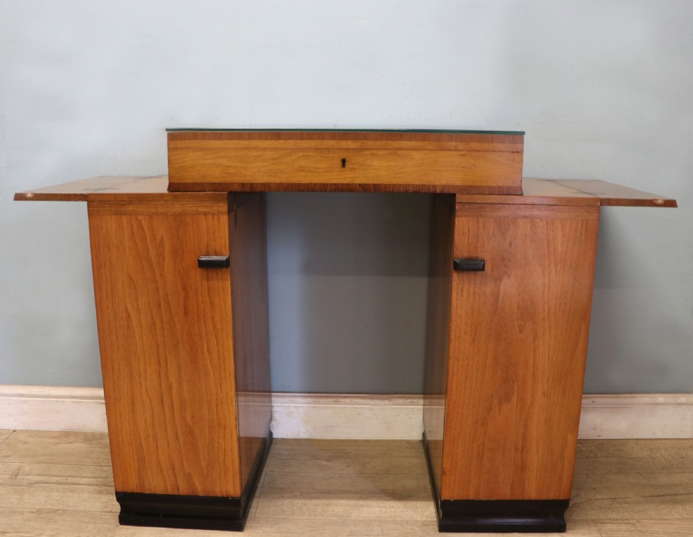 An Art Deco walnut pedestal desk featuring a glass-topped centre writing surface and hinged fold-out side flaps supported by twin cabinet bases. View II