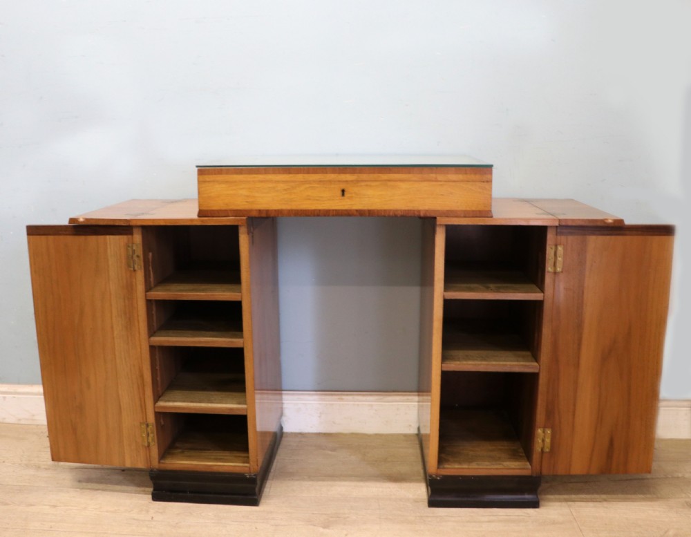 An Art Deco walnut pedestal desk featuring a glass-topped centre writing surface and hinged fold-out side flaps supported by twin cabinet bases. View VII