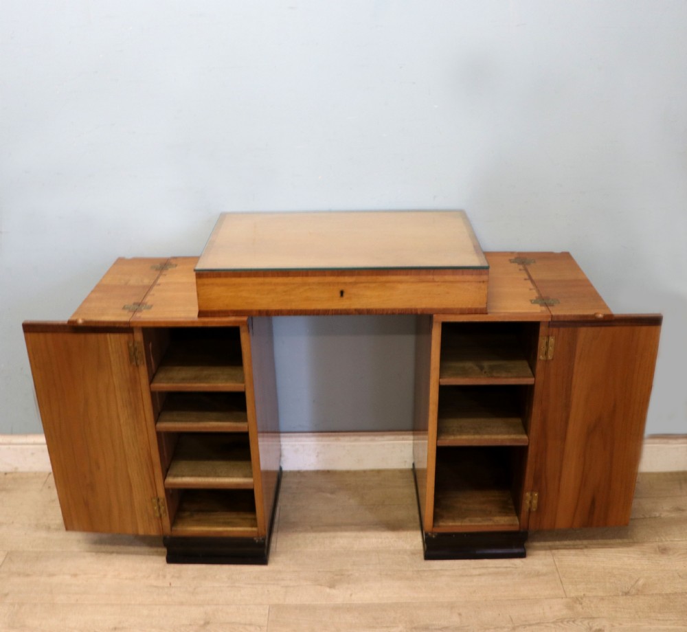 An Art Deco walnut pedestal desk featuring a glass-topped centre writing surface and hinged fold-out side flaps supported by twin cabinet bases. View VIII