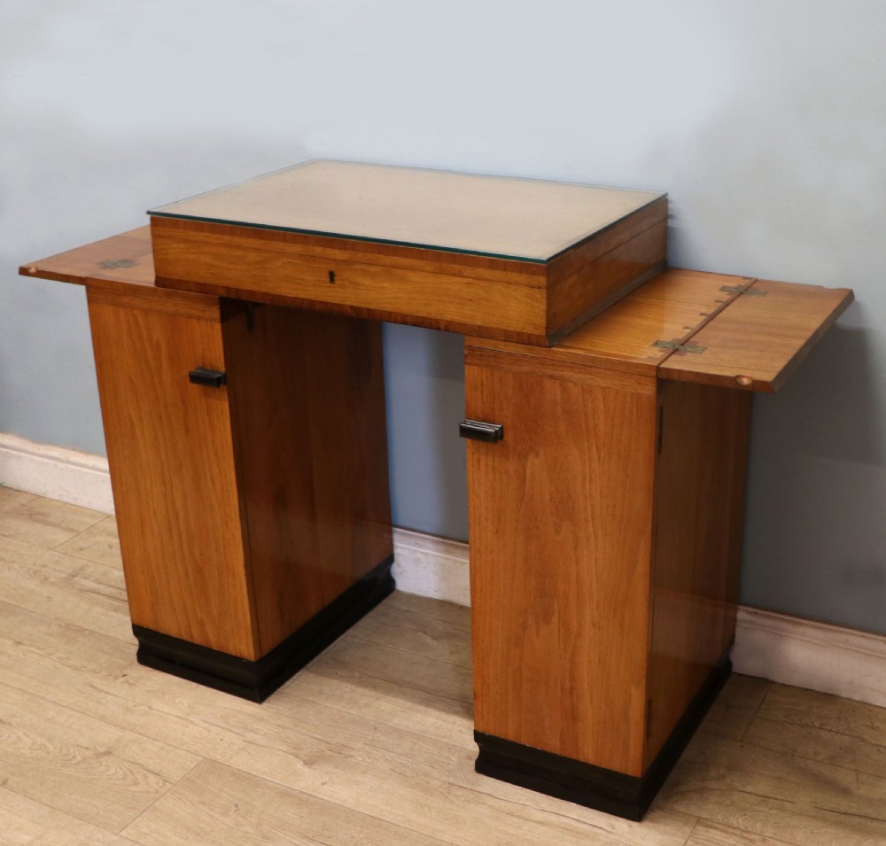 An Art Deco walnut pedestal desk featuring a glass-topped centre writing surface and hinged fold-out side flaps supported by twin cabinet bases. View X