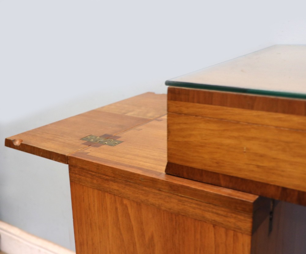 An Art Deco walnut pedestal desk featuring a glass-topped centre writing surface and hinged fold-out side flaps supported by twin cabinet bases. View XV