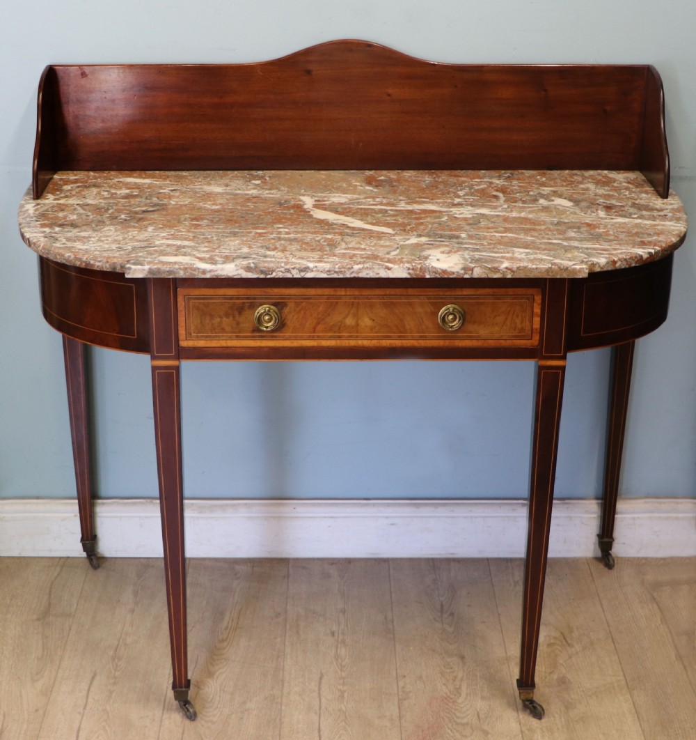 An Edwardian mahogany and satinwood washstand featuring a shaped marble top, wooden splashback, central drawer, and tapered legs on brass castors.