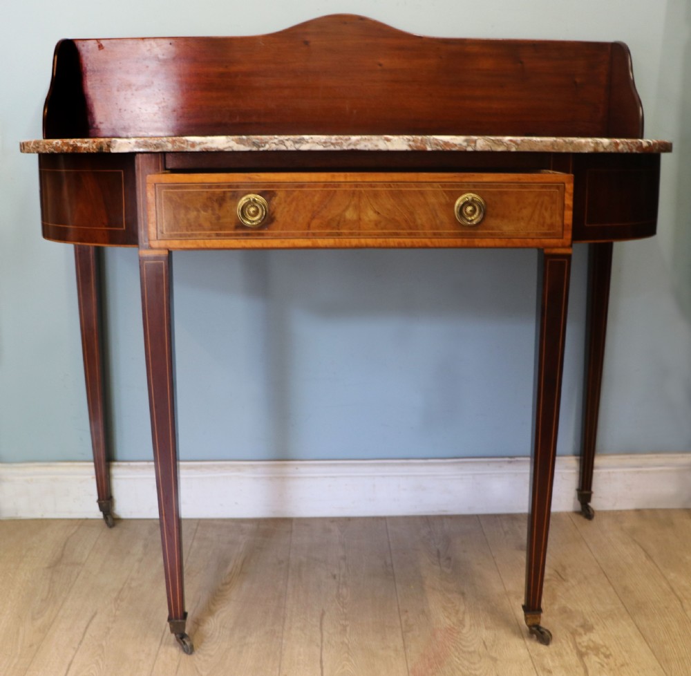 An Edwardian mahogany and satinwood washstand featuring a shaped marble top, wooden splashback, central drawer, and tapered legs on brass castors. View II