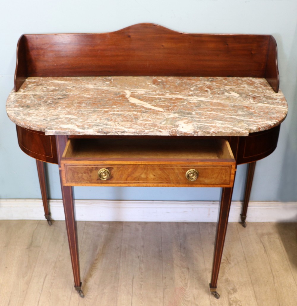 An Edwardian mahogany and satinwood washstand featuring a shaped marble top, wooden splashback, central drawer, and tapered legs on brass castors. View III