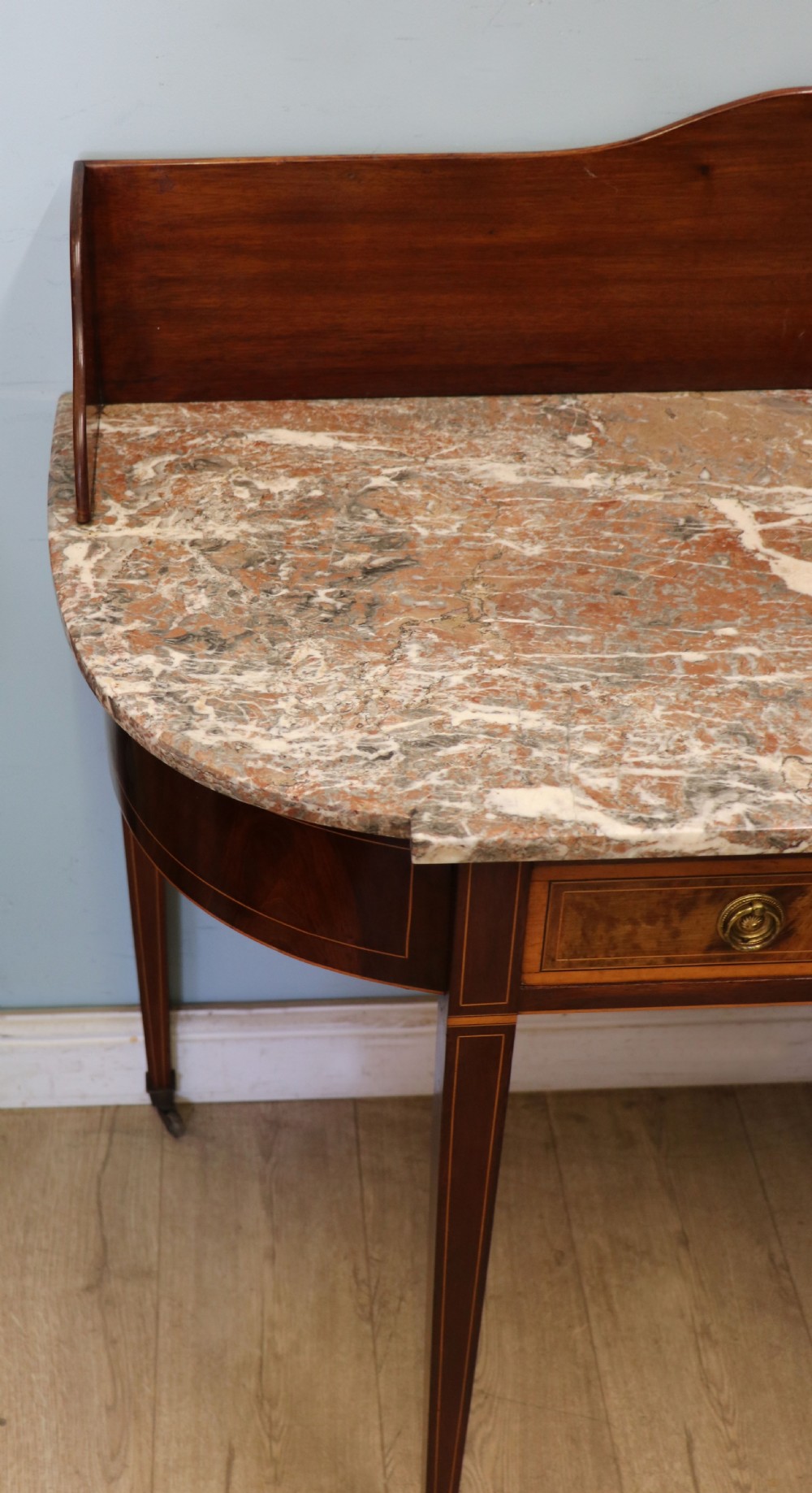 An Edwardian mahogany and satinwood washstand featuring a shaped marble top, wooden splashback, central drawer, and tapered legs on brass castors. View V