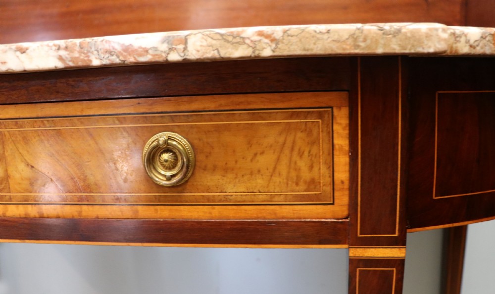 An Edwardian mahogany and satinwood washstand featuring a shaped marble top, wooden splashback, central drawer, and tapered legs on brass castors. View XI