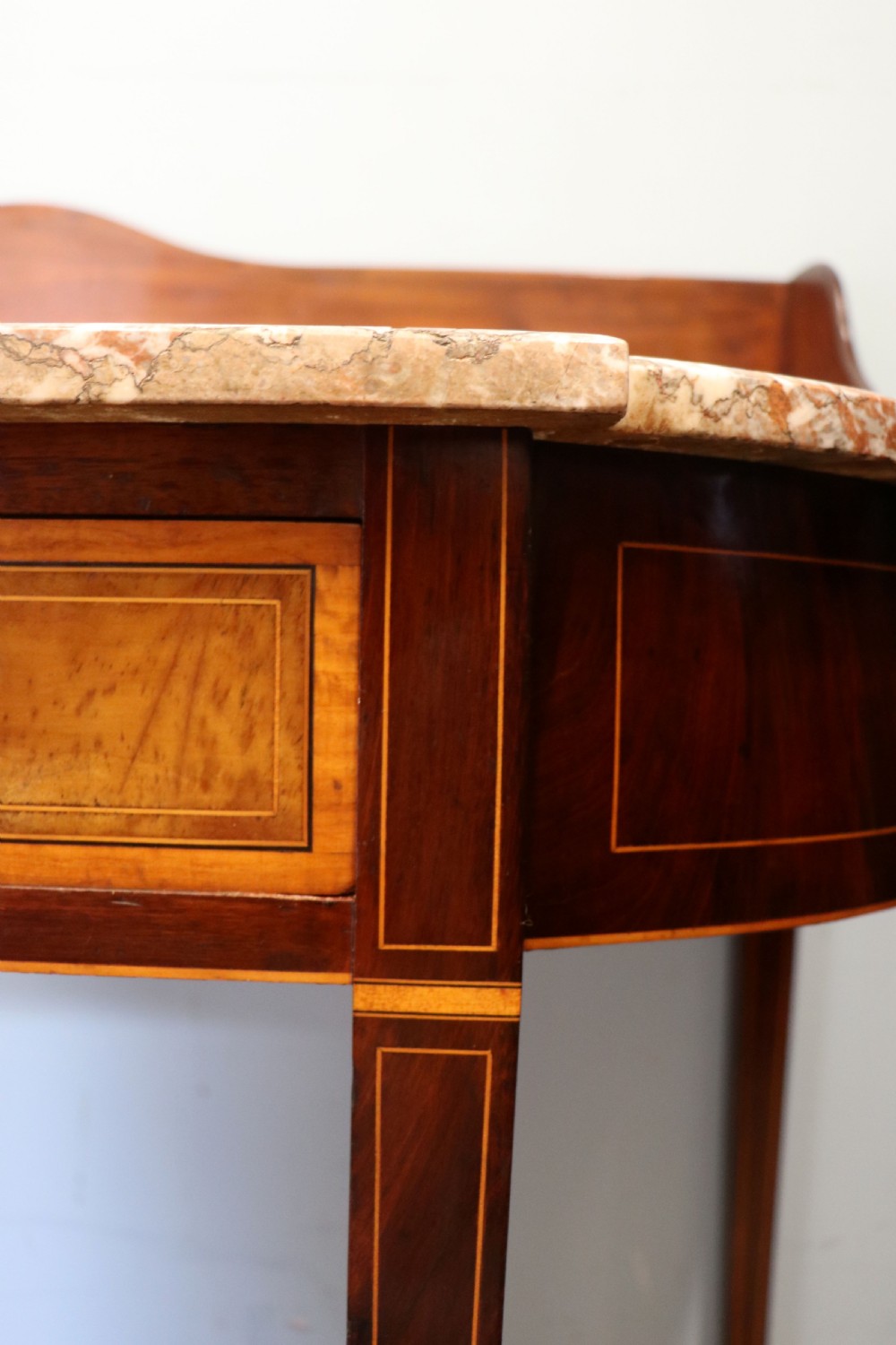 An Edwardian mahogany and satinwood washstand featuring a shaped marble top, wooden splashback, central drawer, and tapered legs on brass castors. View XIII