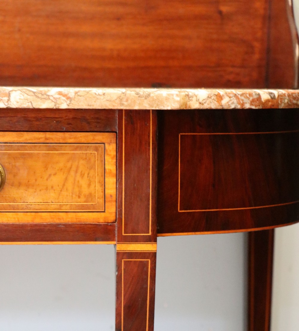 An Edwardian mahogany and satinwood washstand featuring a shaped marble top, wooden splashback, central drawer, and tapered legs on brass castors. View XV
