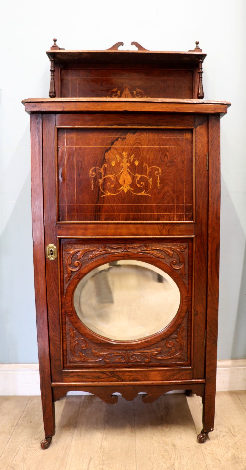 A late nineteenth-century dark hardwood music cabinet featuring floral marquetry inlay on the upper door, an oval mirror panel framed by scroll carving below, and a galleried upper shelf.