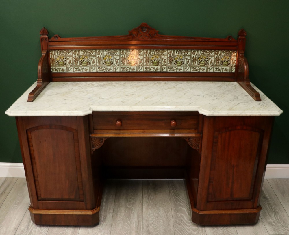 A Victorian Aesthetic Movement mahogany washstand featuring a white marble top, a tiled gallery splashback with floral motifs, two pedestal cupboards, and a central drawer.