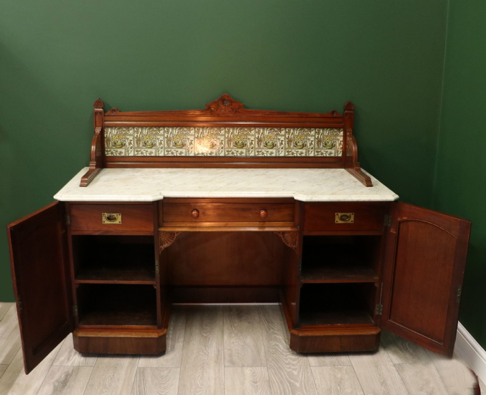 A Victorian Aesthetic Movement mahogany washstand featuring a white marble top, a tiled gallery splashback with floral motifs, two pedestal cupboards, and a central drawer. View IV