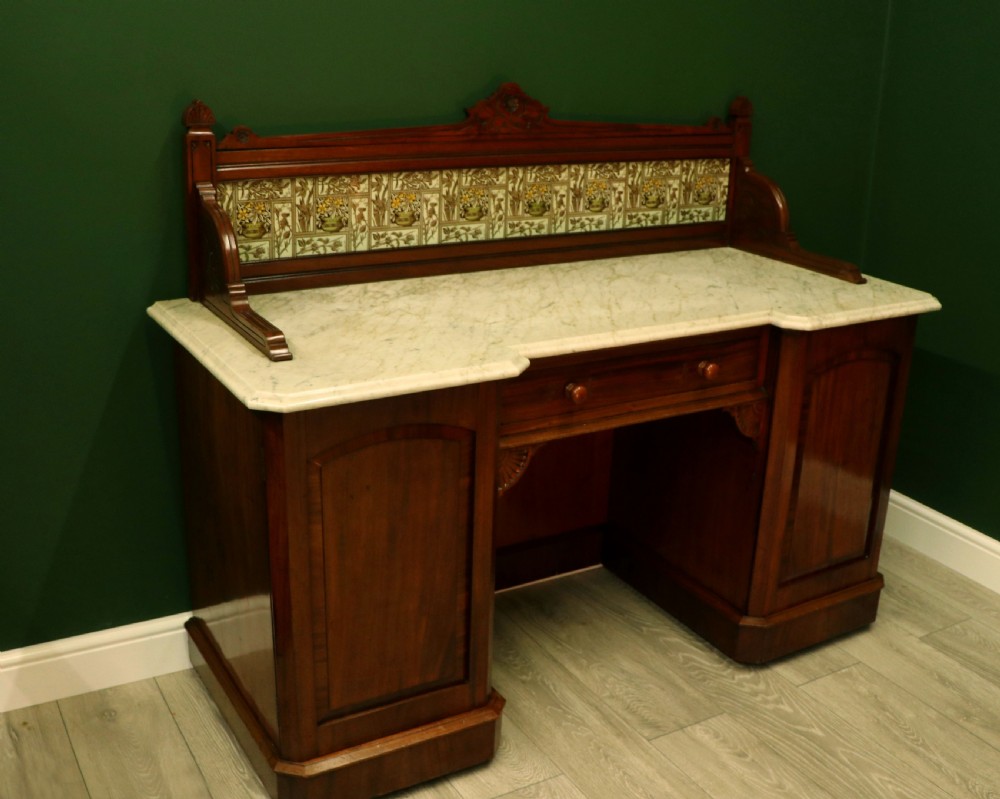 A Victorian Aesthetic Movement mahogany washstand featuring a white marble top, a tiled gallery splashback with floral motifs, two pedestal cupboards, and a central drawer. View V