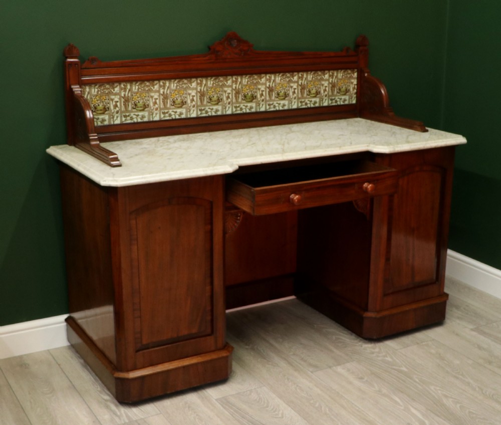 A Victorian Aesthetic Movement mahogany washstand featuring a white marble top, a tiled gallery splashback with floral motifs, two pedestal cupboards, and a central drawer. View VI