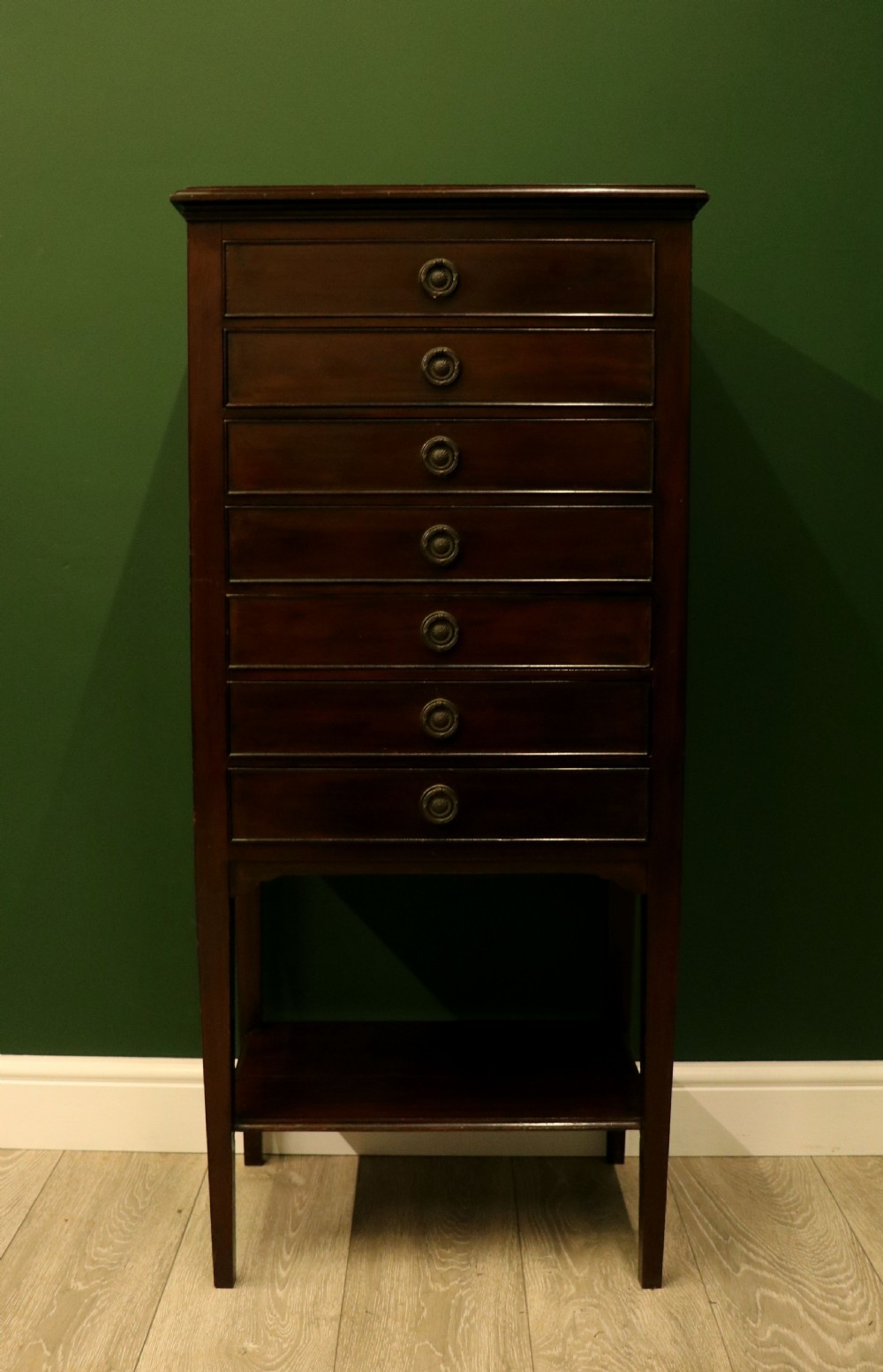 A tall, narrow dark mahogany music cabinet featuring seven fall-front drawers with central brass ring handles and an open shelf near the base.