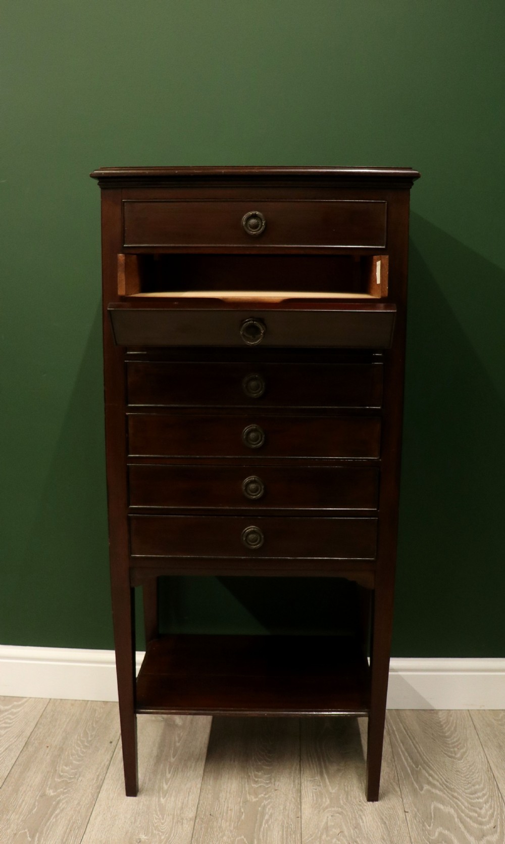 A tall, narrow dark mahogany music cabinet featuring seven fall-front drawers with central brass ring handles and an open shelf near the base. View III