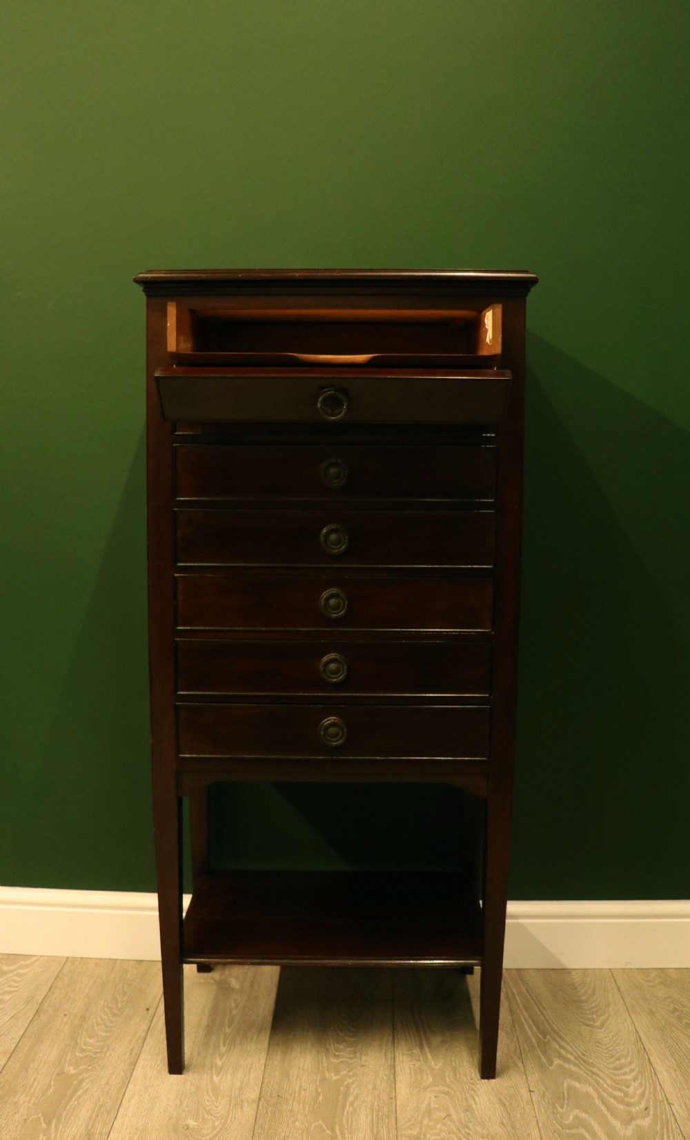 A tall, narrow dark mahogany music cabinet featuring seven fall-front drawers with central brass ring handles and an open shelf near the base. View IV