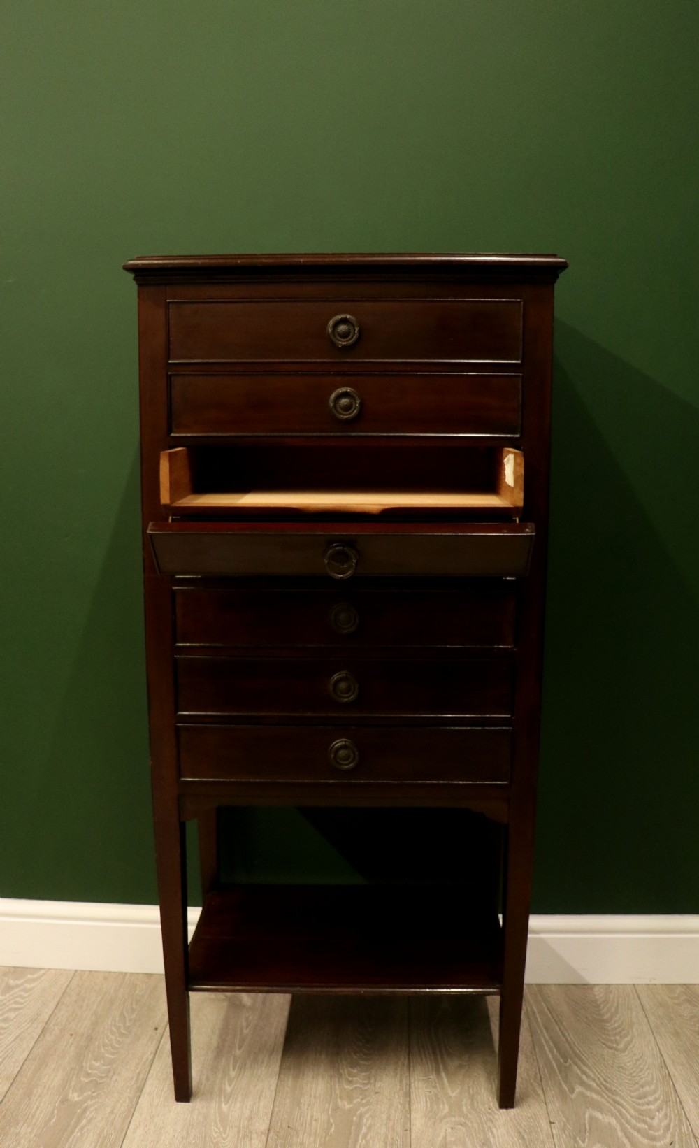 A tall, narrow dark mahogany music cabinet featuring seven fall-front drawers with central brass ring handles and an open shelf near the base. View V