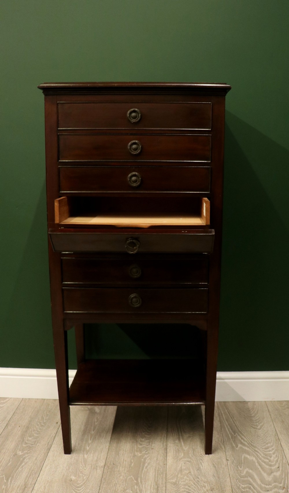 A tall, narrow dark mahogany music cabinet featuring seven fall-front drawers with central brass ring handles and an open shelf near the base. View VI