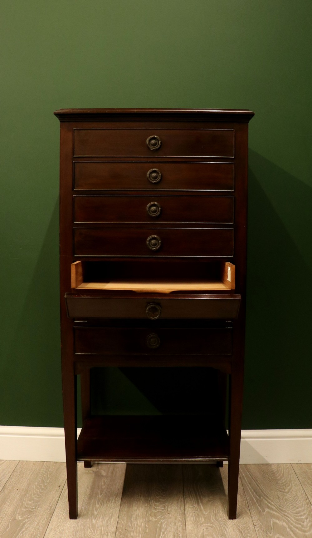 A tall, narrow dark mahogany music cabinet featuring seven fall-front drawers with central brass ring handles and an open shelf near the base. View VII