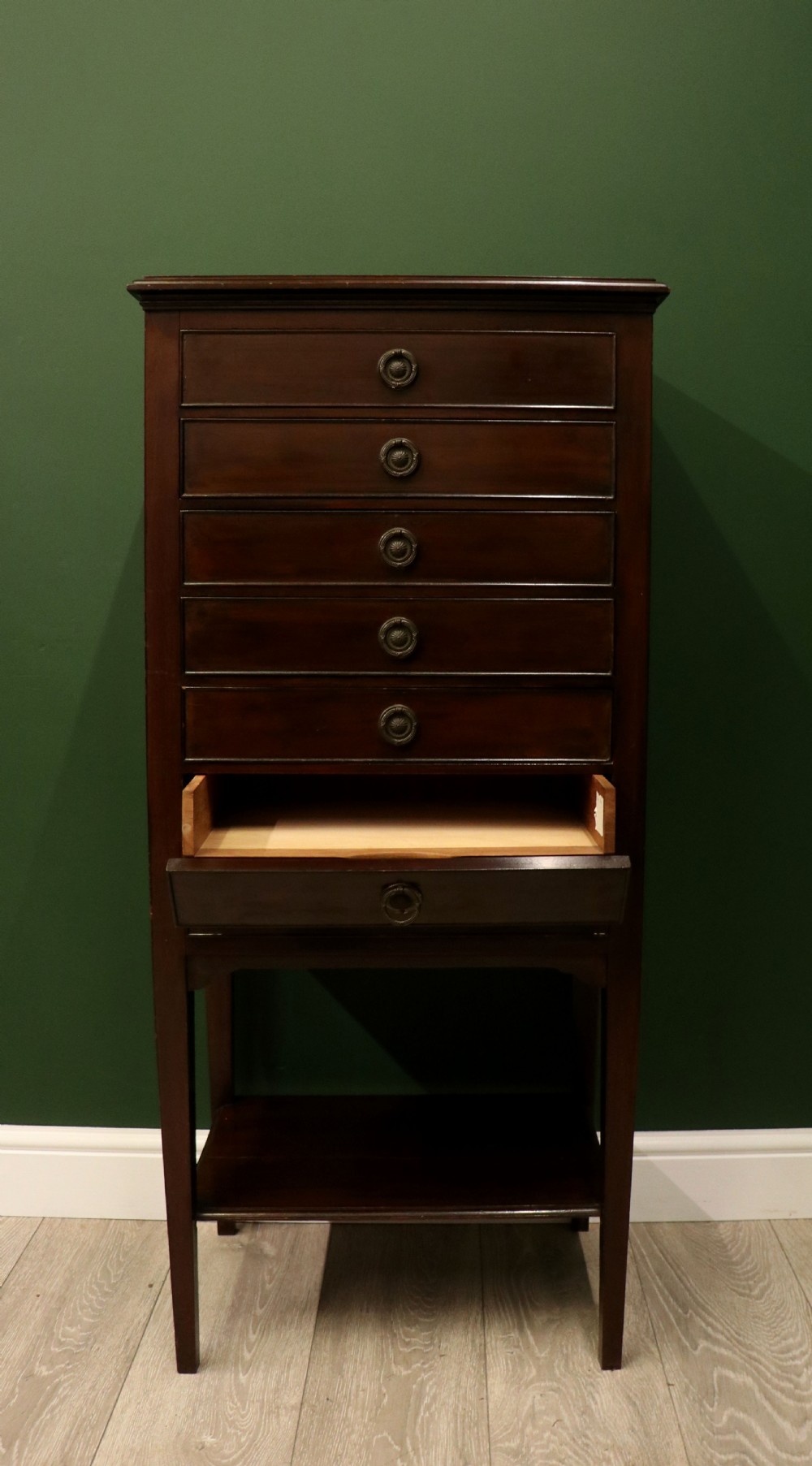 A tall, narrow dark mahogany music cabinet featuring seven fall-front drawers with central brass ring handles and an open shelf near the base. View VIII