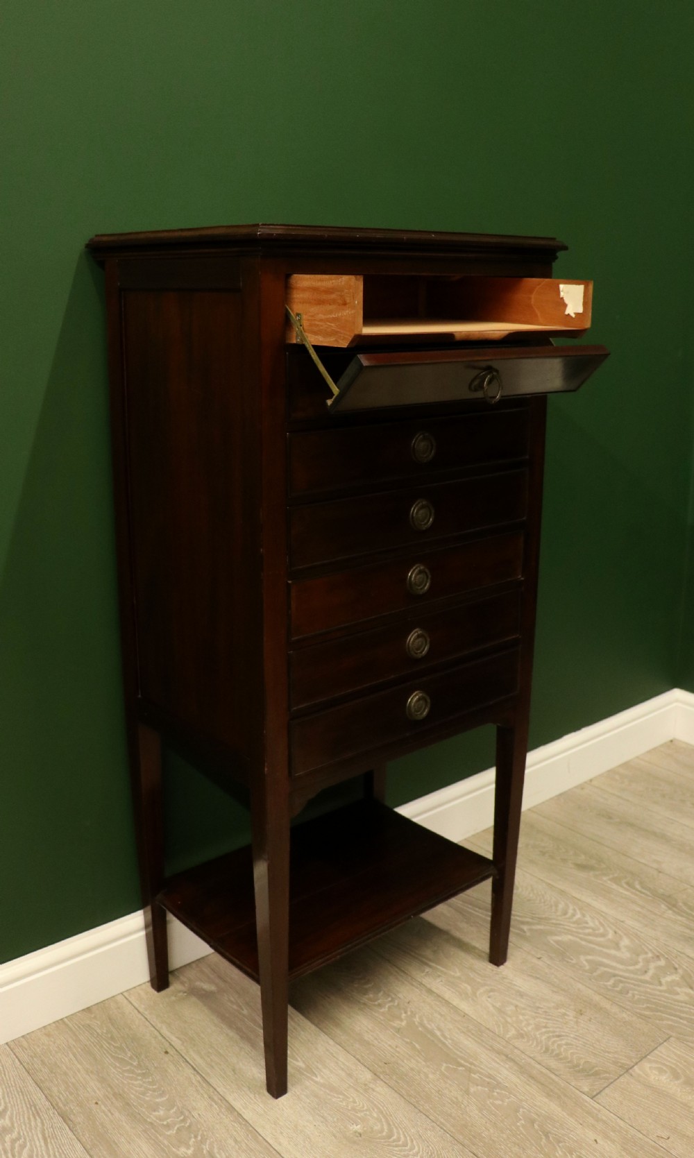 A tall, narrow dark mahogany music cabinet featuring seven fall-front drawers with central brass ring handles and an open shelf near the base. View IX