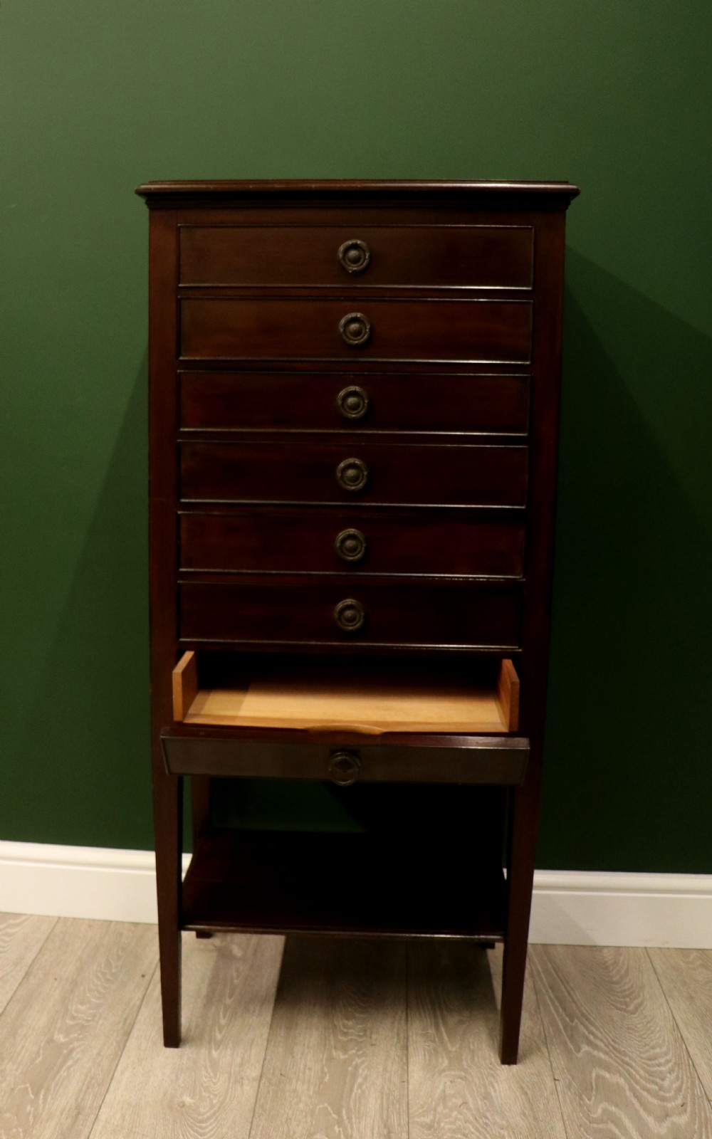 A tall, narrow dark mahogany music cabinet featuring seven fall-front drawers with central brass ring handles and an open shelf near the base. View XI