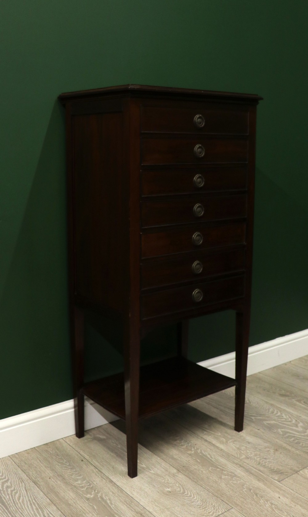A tall, narrow dark mahogany music cabinet featuring seven fall-front drawers with central brass ring handles and an open shelf near the base. View XII