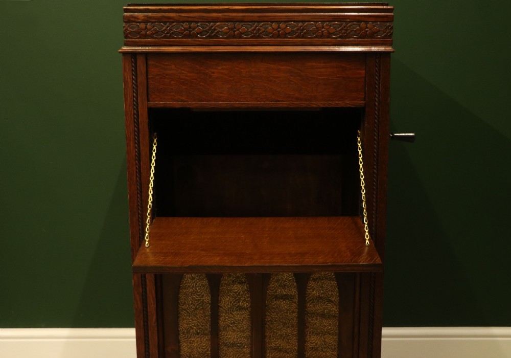 An upright dark oak parlour gramophone cabinet featuring cabriole legs, a hinged lid, carved rope detailing, and gothic arch fretwork grilles. View XXV