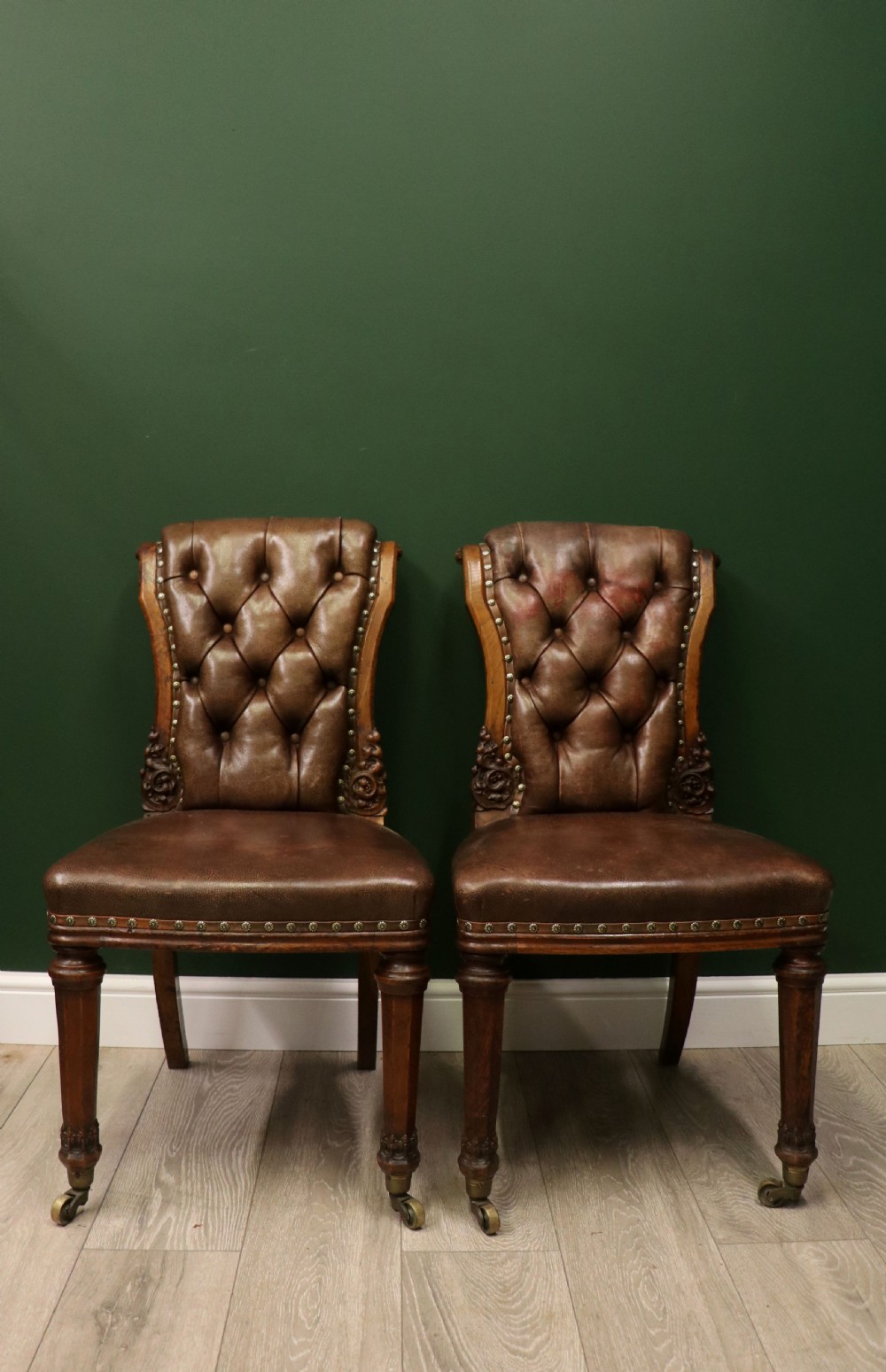 A pair of Victorian carved oak side chairs upholstered in deep-buttoned brown leather with brass studding and turned front legs on castors.