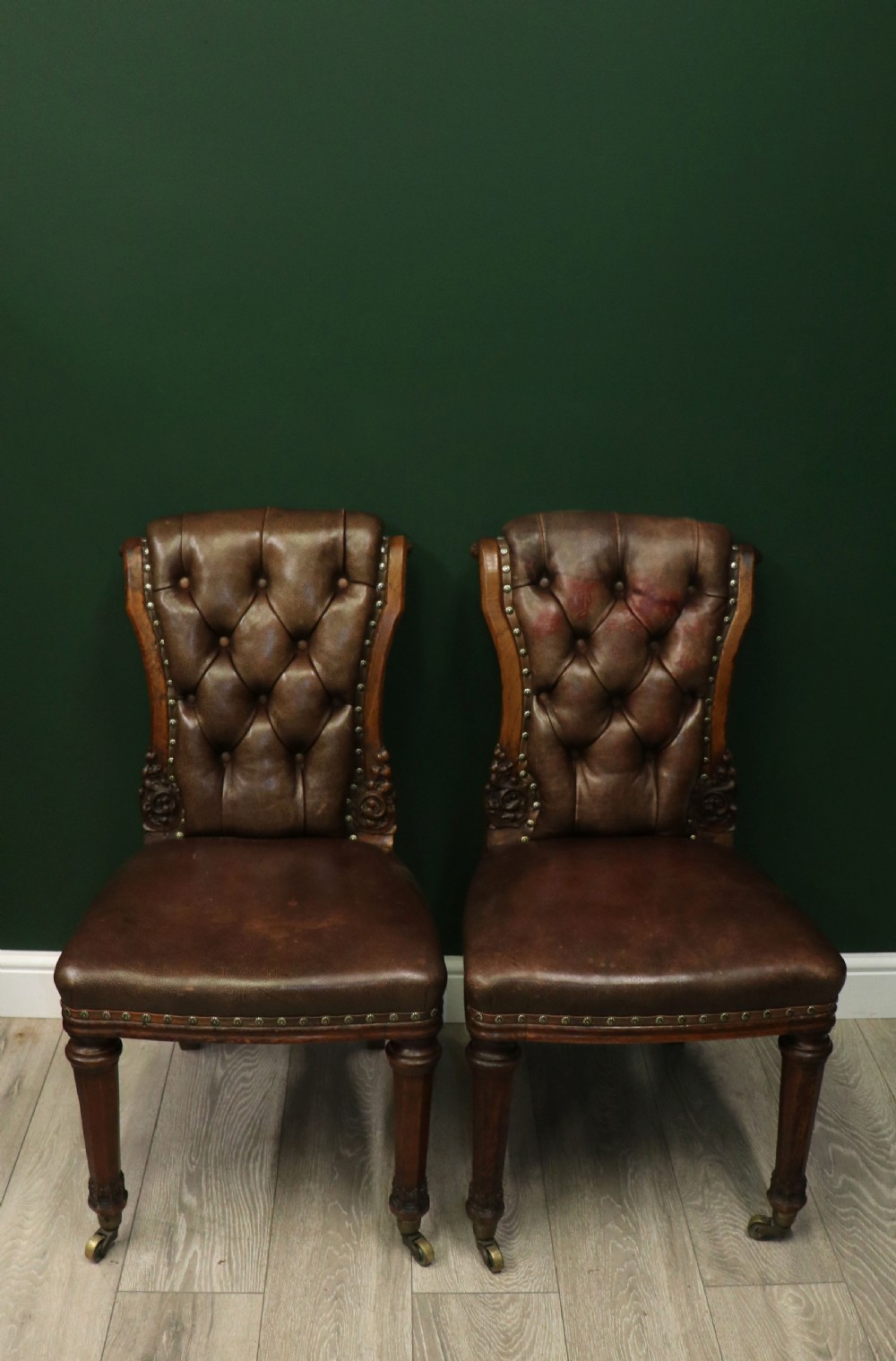 A pair of Victorian carved oak side chairs upholstered in deep-buttoned brown leather with brass studding and turned front legs on castors. View II