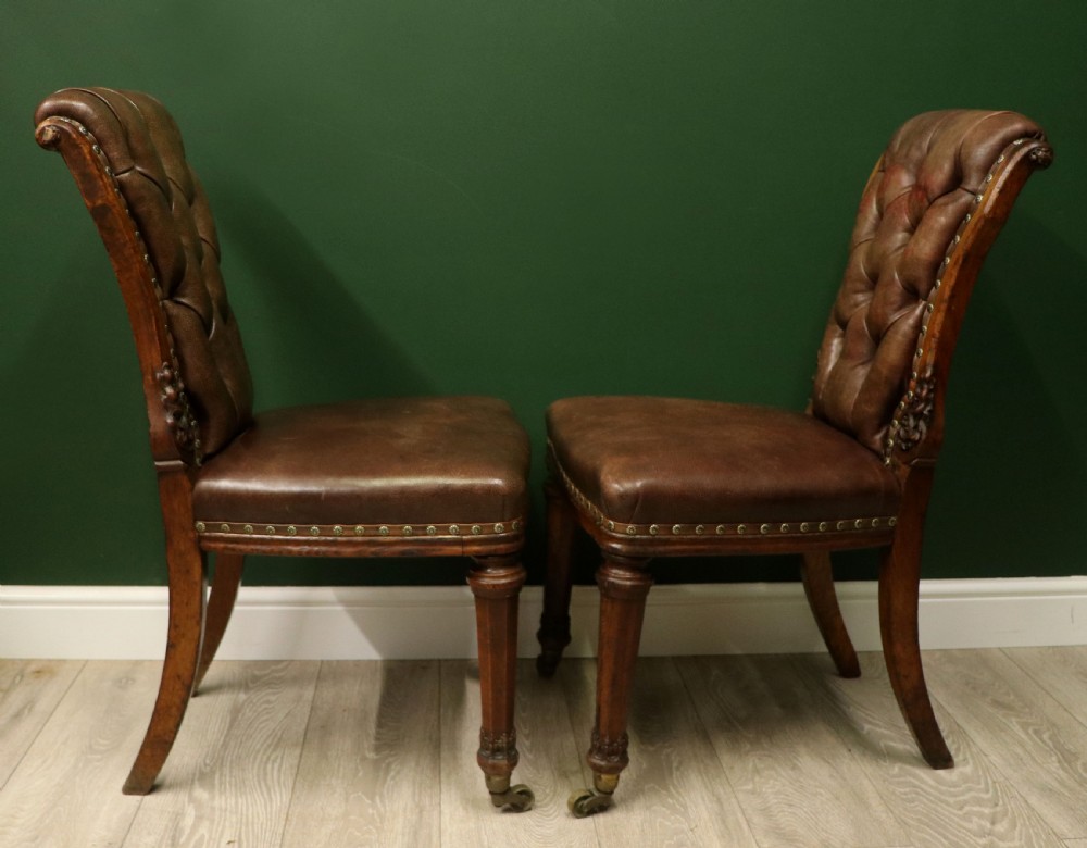 A pair of Victorian carved oak side chairs upholstered in deep-buttoned brown leather with brass studding and turned front legs on castors. View X