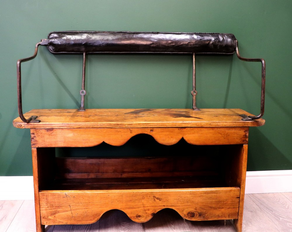 An antique pine wooden bench featuring a shaped aproned base, integrated lower shelf, wrought iron armrests, and a black leather padded cylindrical bolster backrest.