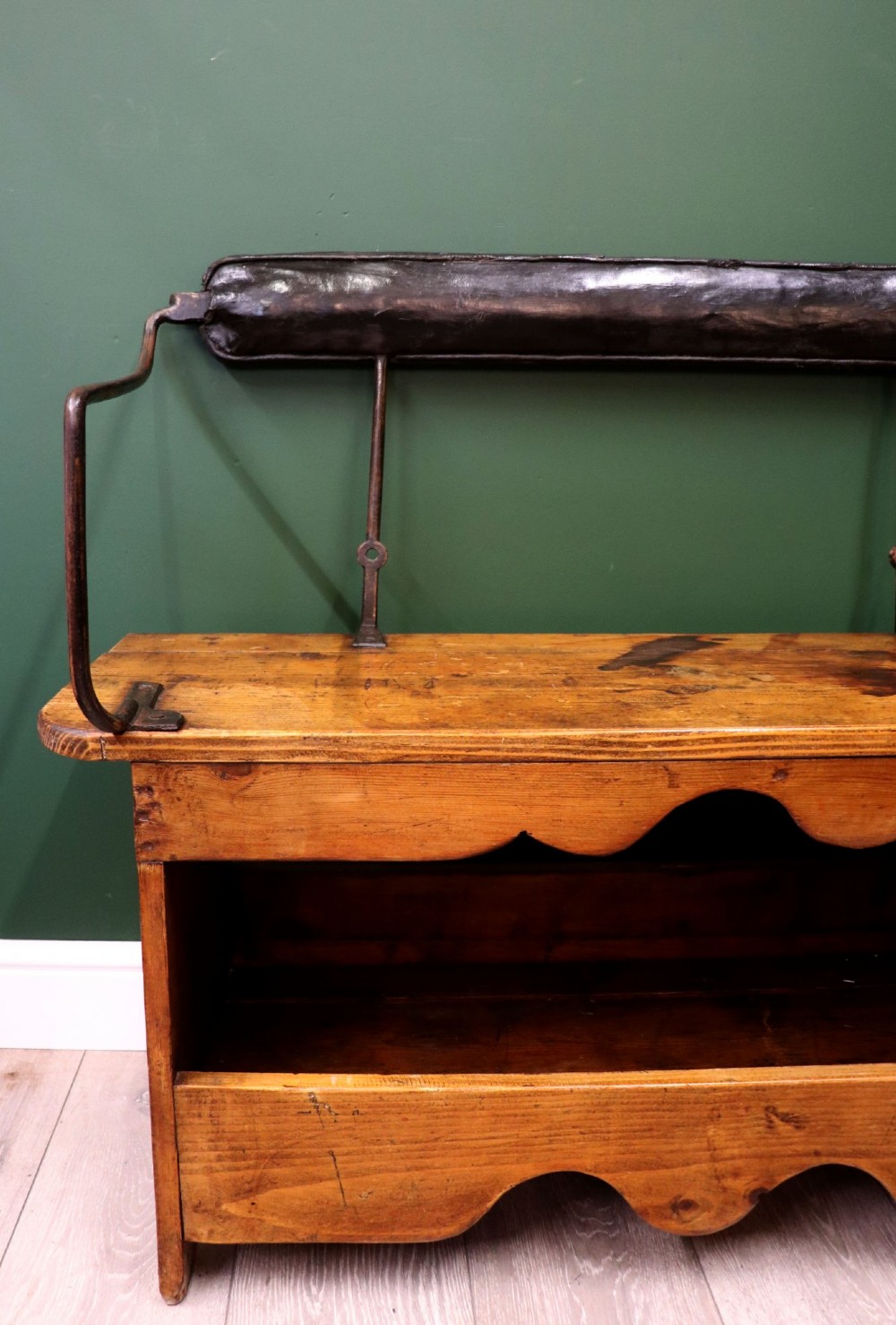 An antique pine wooden bench featuring a shaped aproned base, integrated lower shelf, wrought iron armrests, and a black leather padded cylindrical bolster backrest. View XI