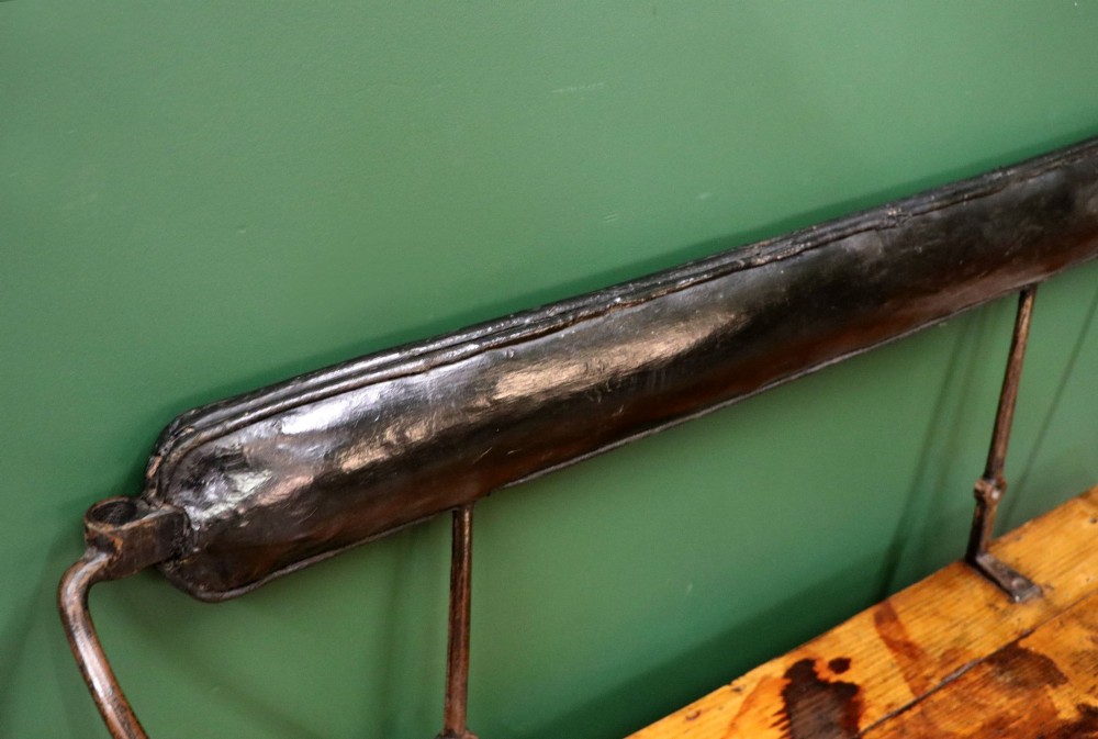 An antique pine wooden bench featuring a shaped aproned base, integrated lower shelf, wrought iron armrests, and a black leather padded cylindrical bolster backrest. View XII