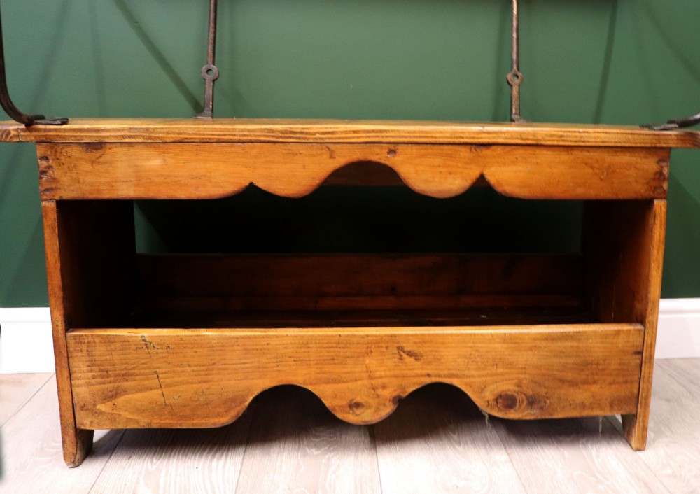 An antique pine wooden bench featuring a shaped aproned base, integrated lower shelf, wrought iron armrests, and a black leather padded cylindrical bolster backrest. View XV