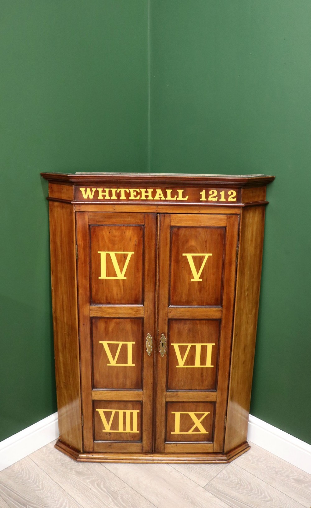 A George III mahogany wall-mounted corner cabinet with double panelled doors featuring painted yellow Roman numerals and a top frieze reading WHITEHALL 1212. View II
