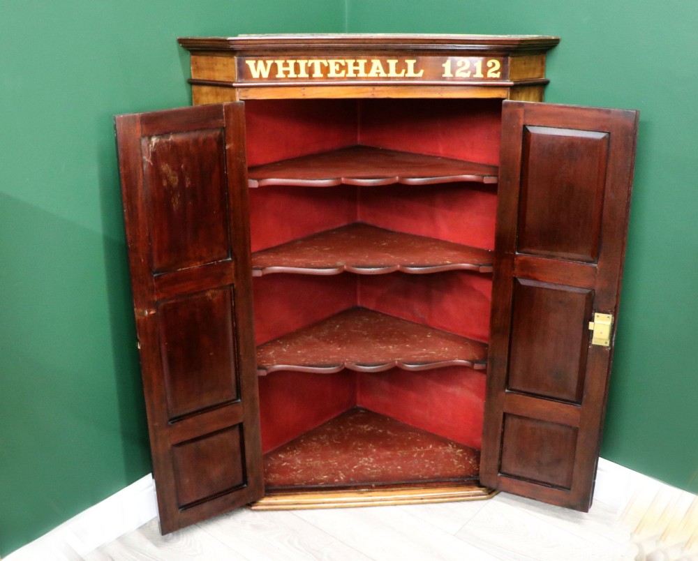 A George III mahogany wall-mounted corner cabinet with double panelled doors featuring painted yellow Roman numerals and a top frieze reading WHITEHALL 1212. View IV