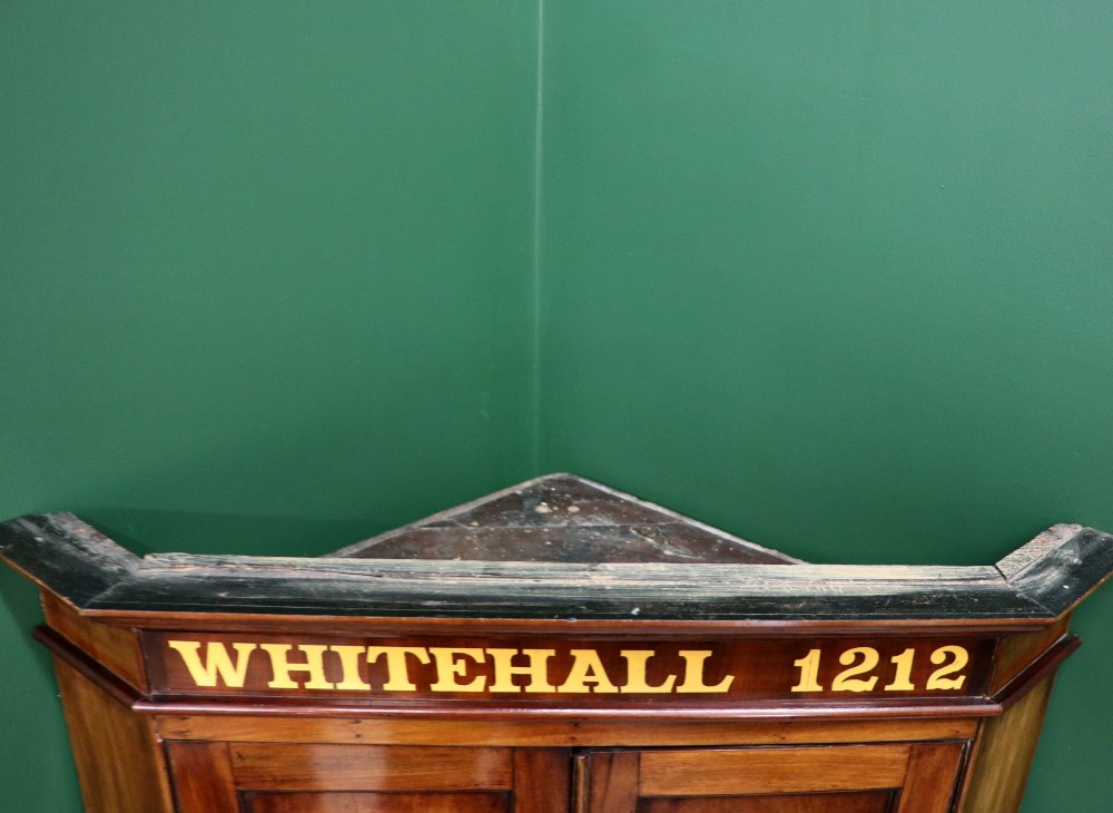 A George III mahogany wall-mounted corner cabinet with double panelled doors featuring painted yellow Roman numerals and a top frieze reading WHITEHALL 1212. View V