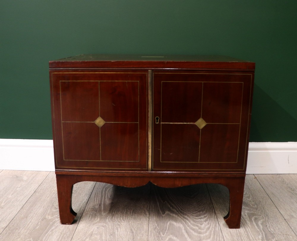 A mahogany campaign style collectors cabinet featuring geometric brass line inlays, twin front doors, a brass central keyhole, and shaped feet.