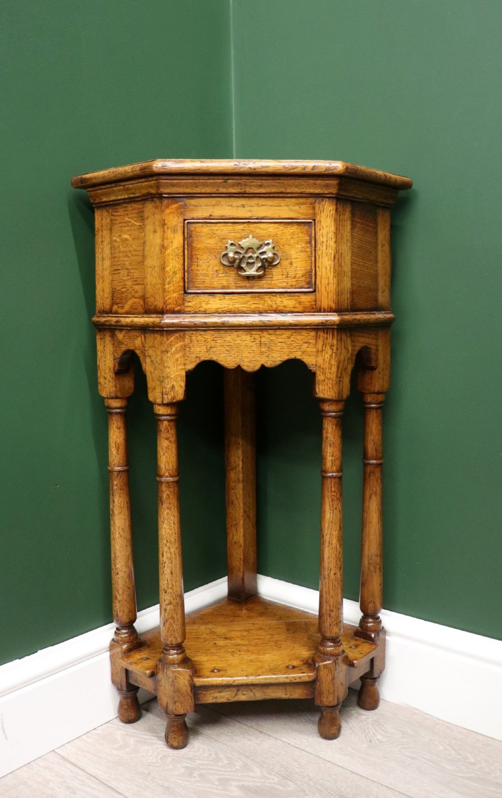 An antique golden oak corner table featuring turned legs, a single drawer with a brass drop handle, a shaped frieze, and a low tier shelf.