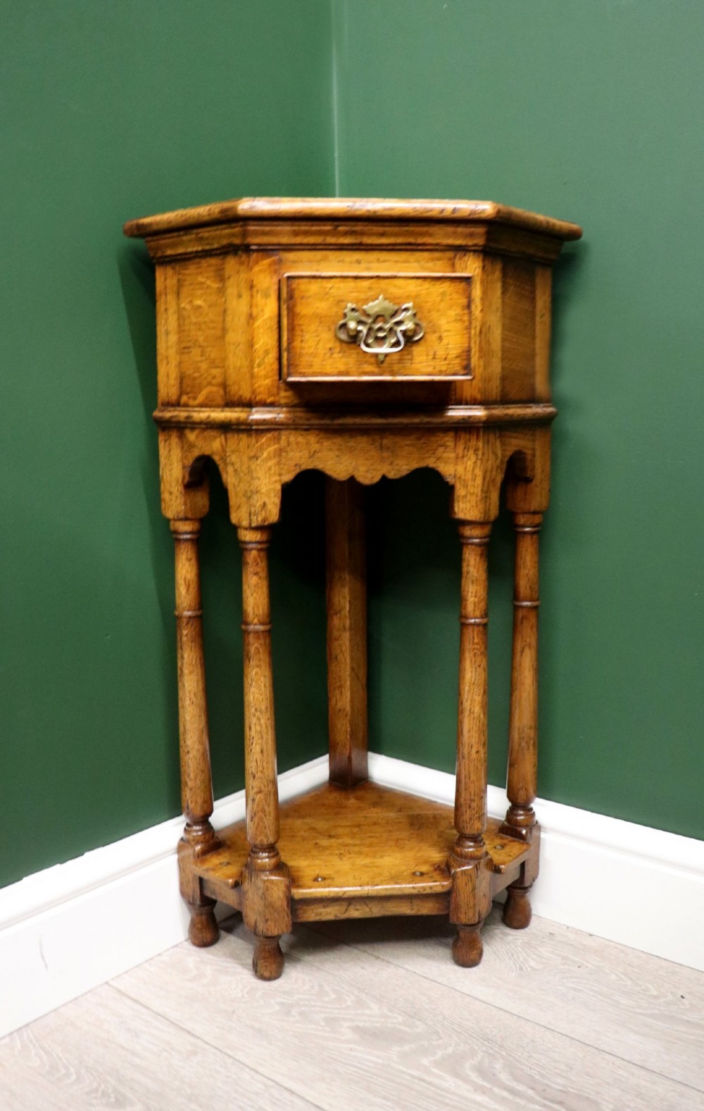 An antique golden oak corner table featuring turned legs, a single drawer with a brass drop handle, a shaped frieze, and a low tier shelf. View II