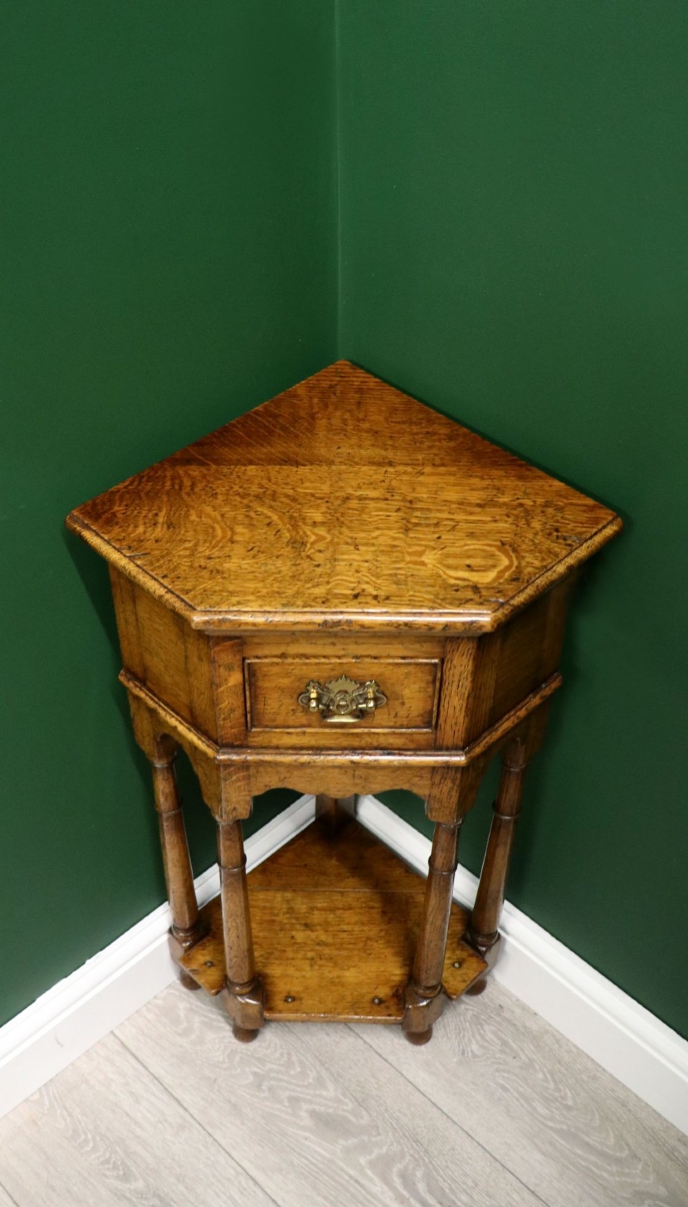 An antique golden oak corner table featuring turned legs, a single drawer with a brass drop handle, a shaped frieze, and a low tier shelf. View III