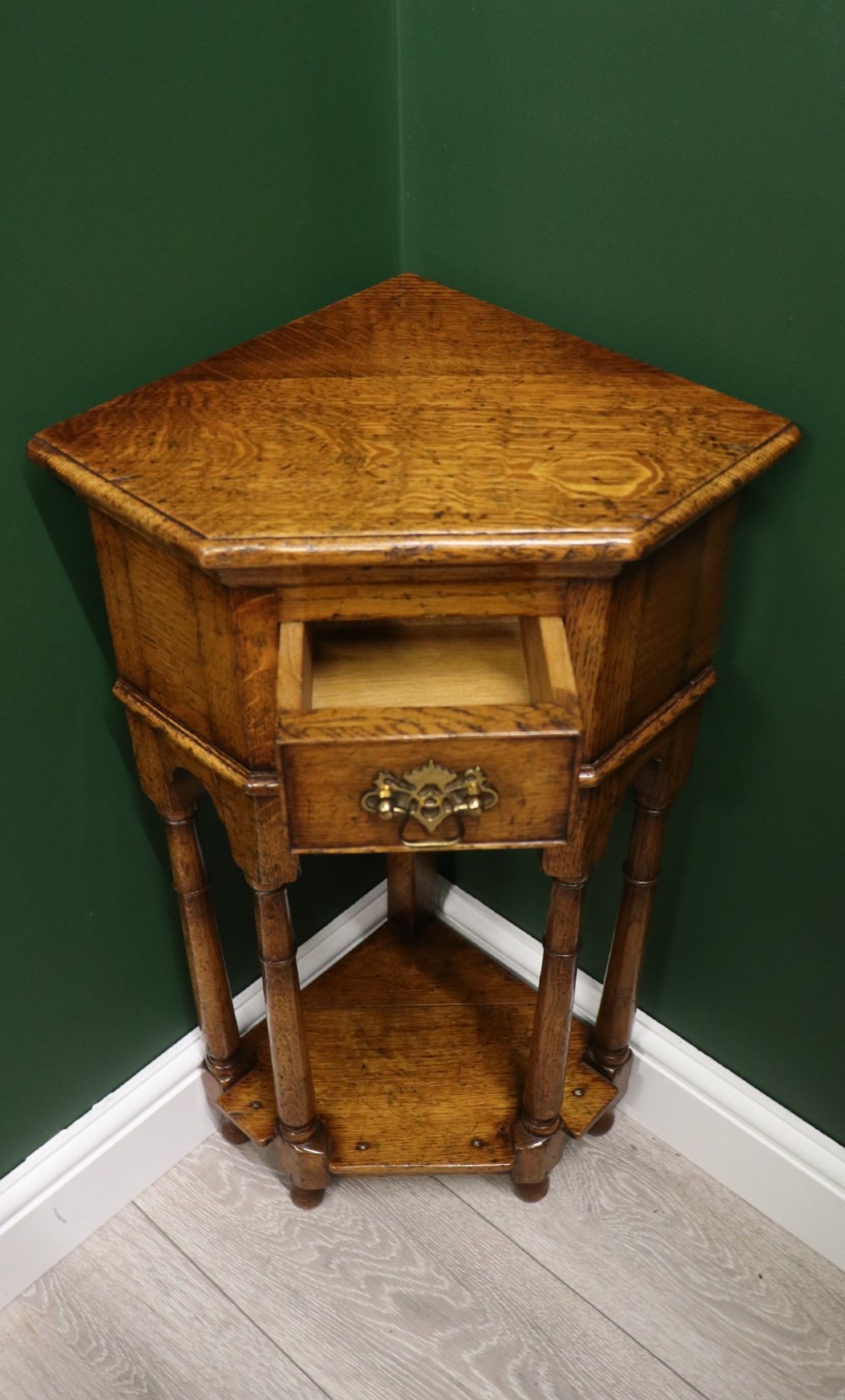 An antique golden oak corner table featuring turned legs, a single drawer with a brass drop handle, a shaped frieze, and a low tier shelf. View IV