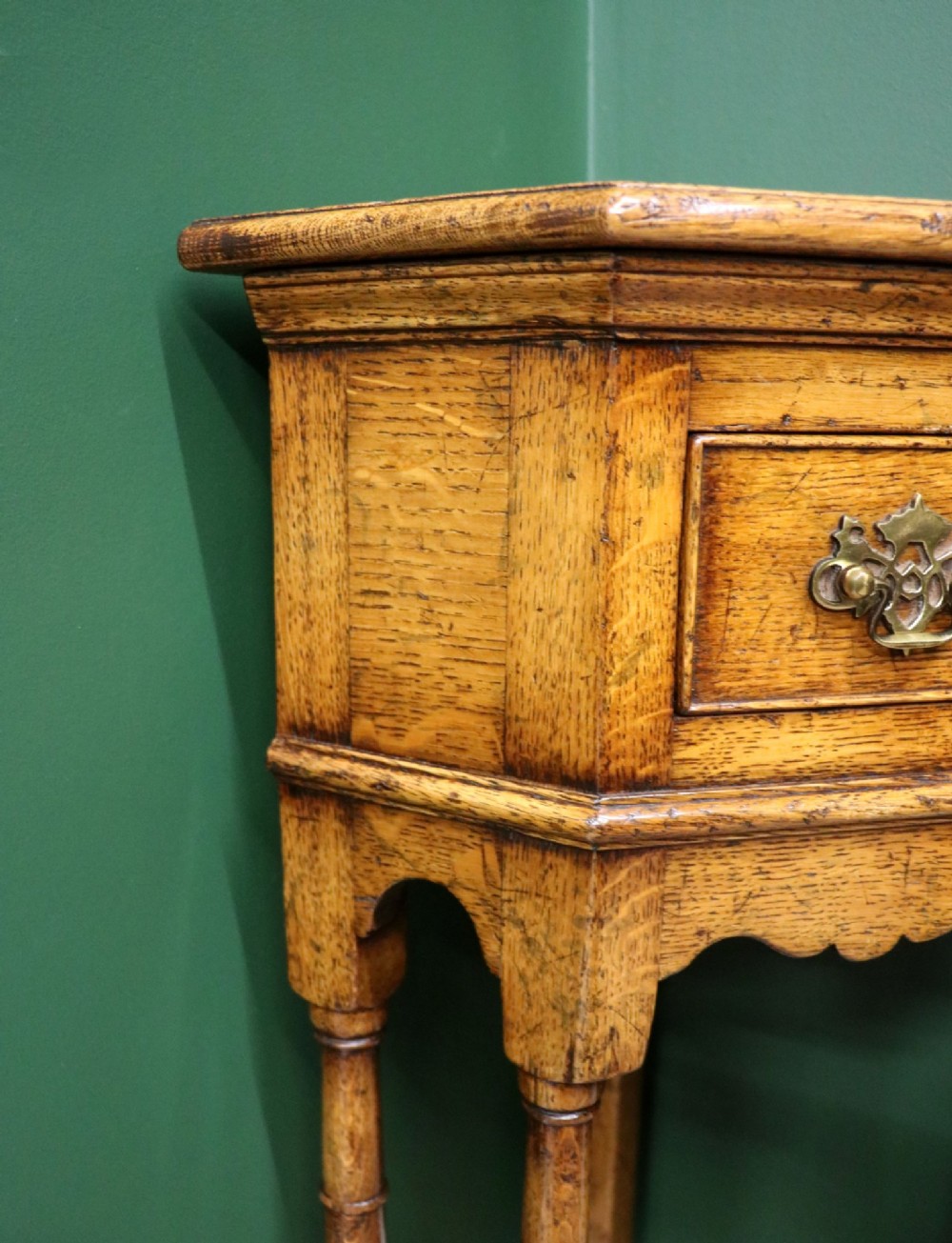 An antique golden oak corner table featuring turned legs, a single drawer with a brass drop handle, a shaped frieze, and a low tier shelf. View XII