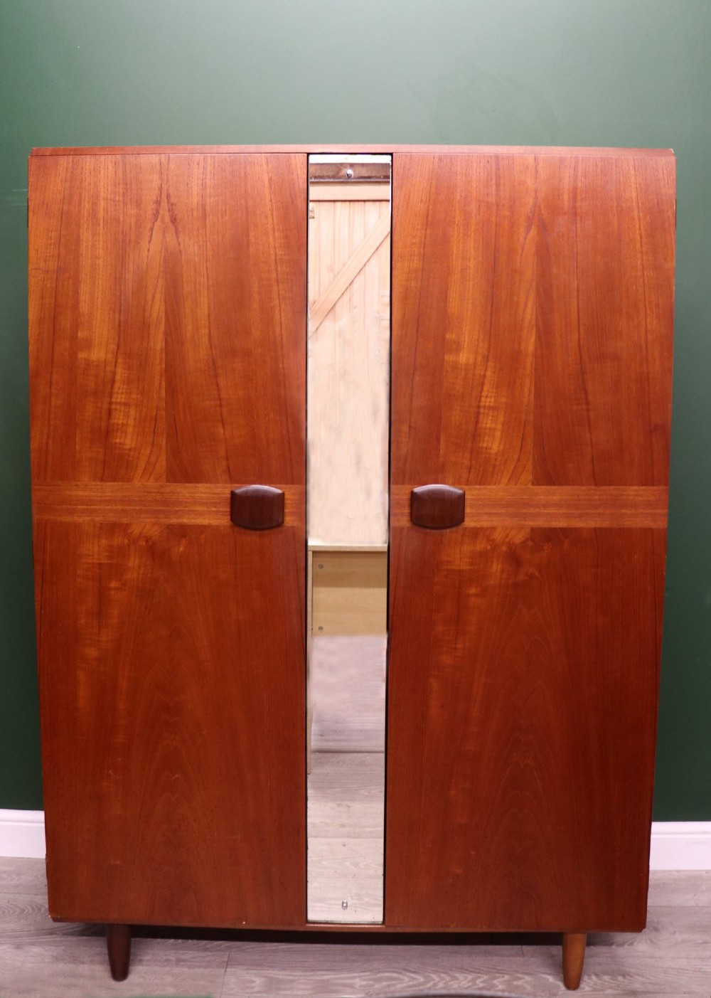 A mid-century teak wood wardrobe featuring two doors with rectangular wooden handles, short tapered legs, and a narrow central full-length mirror.